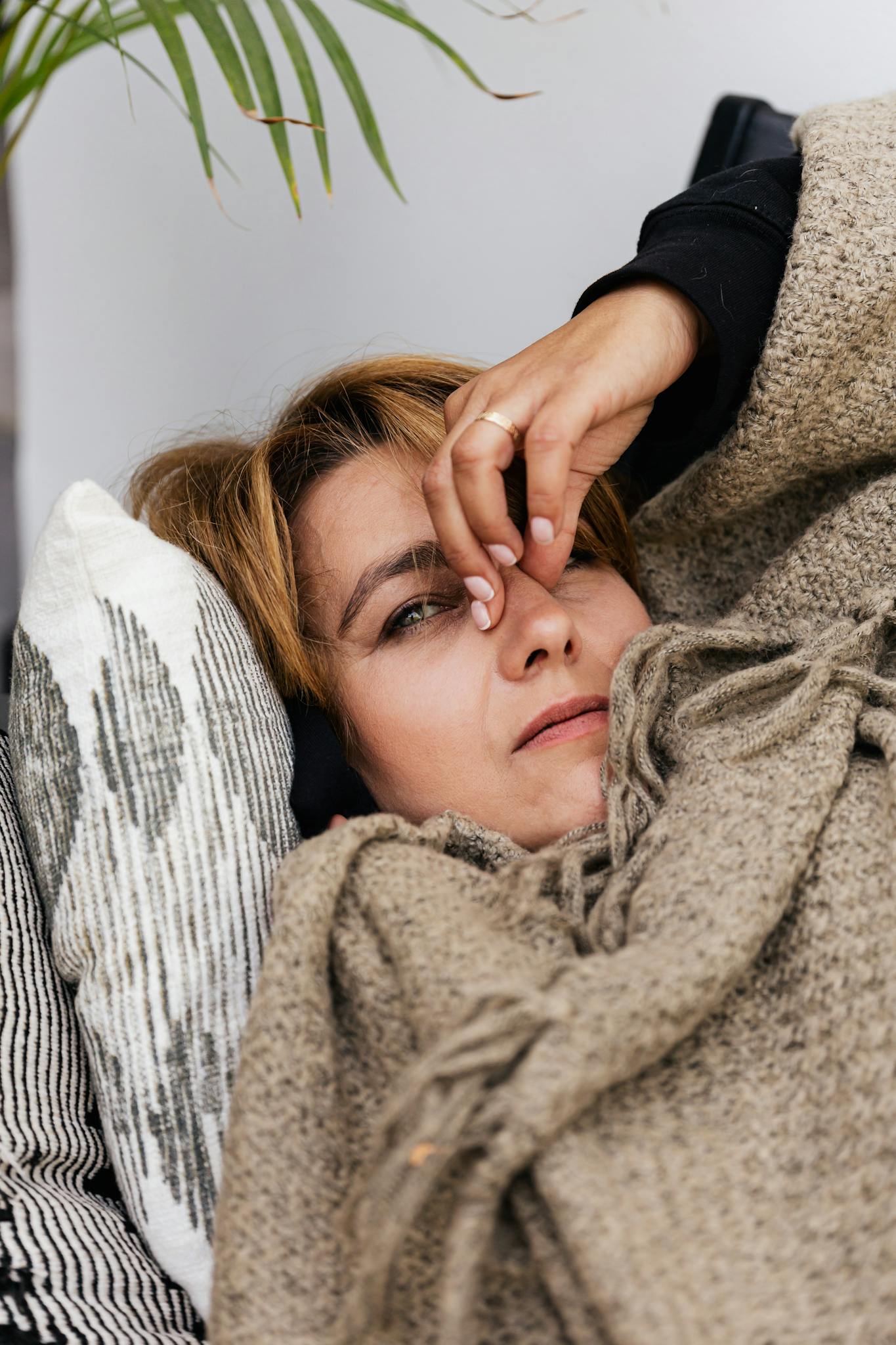 Close-up of a woman resting under a blanket indoors, hand on forehead depicting relaxation or illness.